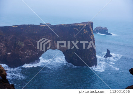 Stone coastline arch on dyrholaey peninsula with nordic foggy landscape and rocky cliffs. Spectacular shoreline stoney gate in iceland, beautiful wilderness with harbor bay view. Stone coastline arch on dyrholaey peninsula with nordic foggy landscape and rocky cliffs. Spectacular shoreline stoney gate in iceland, beautiful wilderness with harbor bay view. 112031096