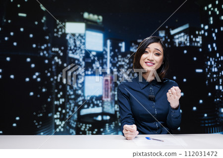Night show host on live broadcast addressing global news topics from newsroom, discussing about daily events. Asian presenter reporting media information to provide latest tv content. 112031472