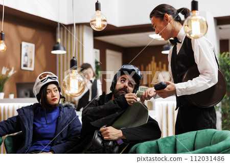 Friendly asian waiter receiving money tip from couple with snow clothing in lounge area of ski mountain resort. Healthy boyfriend and girlfriend thanking employee for good service in winter vacation. 112031486