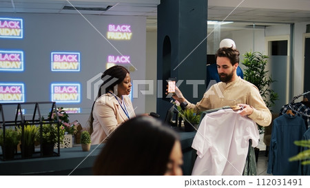 Mobile coupon marketing in retail shop. Client showing smartphone with black friday promo code to cashier, preparing to buy clothes at promotional prices. Shopper using loyalty voucher. 112031491