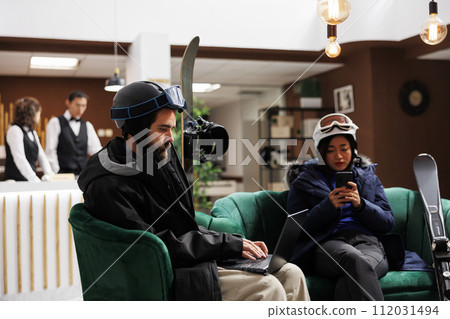 Young couple relaxing in ski resort lounge area with their digital devices enjoying winter holiday. Man with laptop and woman with cell phone taking break from skiing and snowboarding activities. Young couple relaxing in ski resort lounge area with their digital devices enjoying winter holiday. Man with laptop and woman with cell phone taking break from skiing and snowboarding activities. 112031494