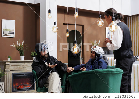 Eager couple outfitted for skiing and snowboarding gear assisted by staff member in hotel lobby. Guests eagerly embrace winter adventure prospects at ski resort set to engage in holiday activities. 112031503