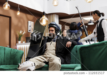 Caucasian man in hotel reception seated in cozy armchair checking his snowboarding equipment. In background bellboy helping female traveler upon arrival at front desk of luxury winter resort. Caucasian man in hotel reception seated in cozy armchair checking his snowboarding equipment. In background bellboy helping female traveler upon arrival at front desk of luxury winter resort. 112031517