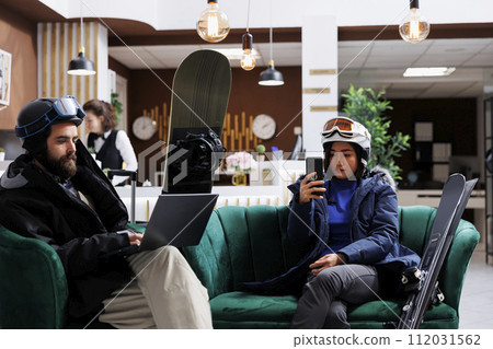 Young couple enjoys winter vacation by lounging in the ski resort lounge area with their digital gadgets. A man with a laptop and a lady with smartphone take a break from skiing and snowboarding. Young couple enjoys winter vacation by lounging in the ski resort lounge area with their digital gadgets. A man with a laptop and a lady with smartphone take a break from skiing and snowboarding. 112031562