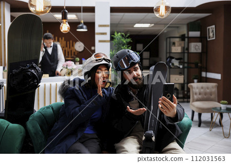 Beautiful couple using smartphone for video call while in lounge area of winter resort. Boyfriend and girlfriend seated with skiing and snowboarding gear engaged in a videoconference on mobile device. Beautiful couple using smartphone for video call while in lounge area of winter resort. Boyfriend and girlfriend seated with skiing and snowboarding gear engaged in a videoconference on mobile device. 112031563