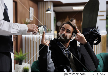 Close-up view showcasing caucasian male tourist with snowboarding gear giving cash tip to employee of exclusive winter mountain resort. Detailed shot of young man tipping ski hotel concierge. Close-up view showcasing caucasian male tourist with snowboarding gear giving cash tip to employee of exclusive winter mountain resort. Detailed shot of young man tipping ski hotel concierge. 112031567