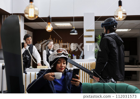 Arriving at ski resort diverse guests check-in, browse on digital devices, and receive top-notch customer service. Skiing and snowboarding gear and technology create modern winter holiday atmosphere. 112031573