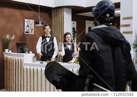 At front desk of exclusive winter resort, two receptionists warmly welcoming male guest in snow gear. Tourist wearing winter jacket and carrying snowboarding equipment arrives in hotel lobby. At front desk of exclusive winter resort, two receptionists warmly welcoming male guest in snow gear. Tourist wearing winter jacket and carrying snowboarding equipment arrives in hotel lobby. 112031582