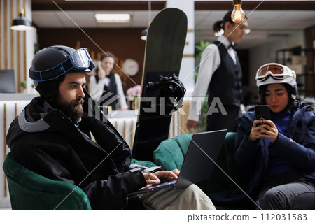 Two visitors sit comfortably using digital devices while snowboard equipment suggests nearby winter sports. Image showing man and woman surfing on laptop and mobile phone in ski hotel lobby. 112031583
