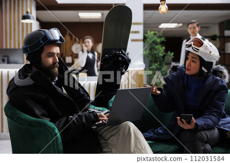 In hotel lobby young guests on couch plan skiing and snowboarding activities with digital devices. Couple using technology for online check-in at ski resort. Snowboard helmets and laptop available. In hotel lobby young guests on couch plan skiing and snowboarding activities with digital devices. Couple using technology for online check-in at ski resort. Snowboard helmets and laptop available. 112031584