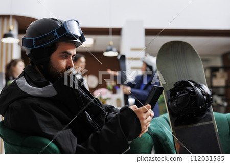 Male tourist in winter attire relaxes on sofa in hotel lobby, browsing on digital tablet. Caucasian man with snowboarding gear makes reservation with smart device for winter holiday at ski resort. Male tourist in winter attire relaxes on sofa in hotel lobby, browsing on digital tablet. Caucasian man with snowboarding gear makes reservation with smart device for winter holiday at ski resort. 112031585
