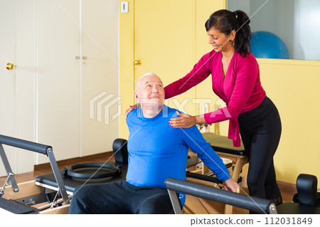 Elderly man practicing pilates on wunda chair under guidance of Hispanic female trainer Elderly man practicing pilates on wunda chair under guidance of Hispanic female trainer 112031849