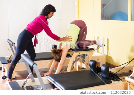 Woman practicing pilates on combo chair supervised by hispanic female trainer Woman practicing pilates on combo chair supervised by hispanic female trainer 112032023