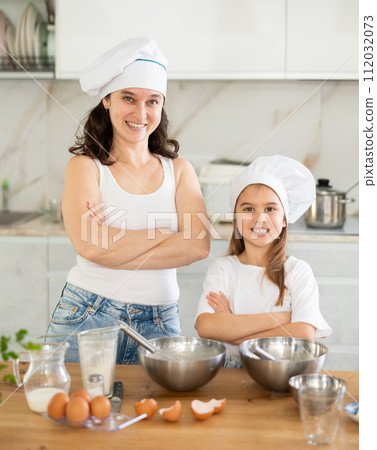 Mother and preteen daughter wearing chef hats standing at table 112032073