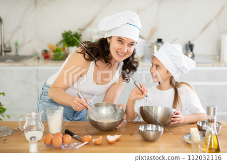 Mom and girl in white chief hats are talking in kitchen and whisking ingredients for pastry in bowl Mom and girl in white chief hats are talking in kitchen and whisking ingredients for pastry in bowl 112032116