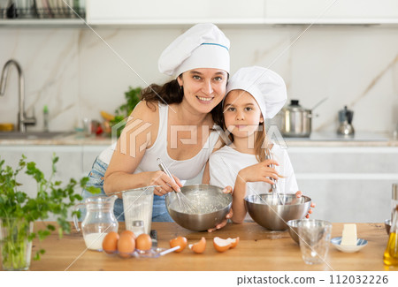 Mom and girl in white chief hats are talking in kitchen and whisking ingredients for pastry in bowl Mom and girl in white chief hats are talking in kitchen and whisking ingredients for pastry in bowl 112032226