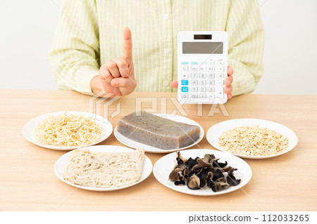 A middle-aged woman holding food containing a lot of dietary fiber and a calculator and raising her index finger 112033265