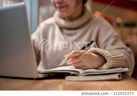 A woman in a comfy sweater is working remotely at a cafe, making list in her notebook. A woman in a comfy sweater is working remotely at a cafe, making list in her notebook. 112033373