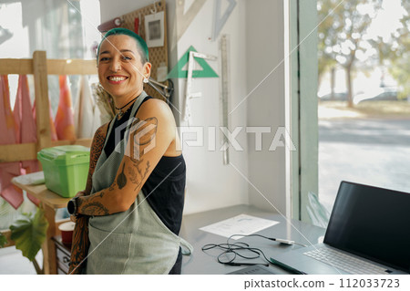 Smiling female designer in apron standing near desk with laptop in own workshop and looks camera Smiling female designer in apron standing near desk with laptop in own workshop and looks camera 112033723