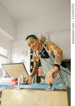 Female worker with squeegee is pressing ink on frame working in design studio. Silk screen printing 112033725