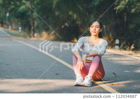 Sport fitness concept, A serene young woman takes a break from running, sitting on a sunlit road surrounded by trees. 112034887