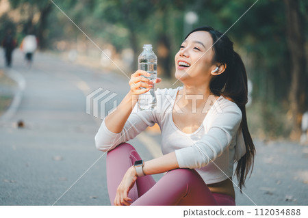 Sport fitness concept, A joyful young woman takes a water break outdoors after exercising, wearing earphones and sportswear, embodying a healthy lifestyle. Sport fitness concept, A joyful young woman takes a water break outdoors after exercising, wearing earphones and sportswear, embodying a healthy lifestyle. 112034888