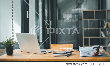 A tidy office desk displaying an open laptop, a small potted plant, and a stack of documents with colorful tabs in a well-lit workspace with bookshelves. 112034908