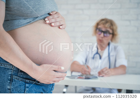Doctor obstetrician gynecologist at his desk in the background. Close-up of a pregnant woman's belly. Doctor obstetrician gynecologist at his desk in the background. Close-up of a pregnant woman's belly. 112035320