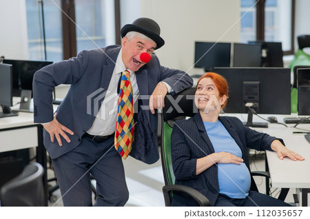 Elderly Caucasian man in clown costume amuses pregnant red-haired woman at work desk in office. Elderly Caucasian man in clown costume amuses pregnant red-haired woman at work desk in office. 112035657