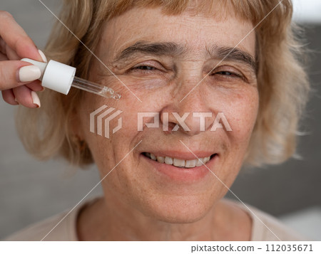Close-up portrait of an old woman applying hyaluronic acid serum with a pipette. Anti-aging face care.  112035671