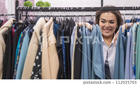 A smiling fat woman in a plus size store chooses clothes while sorting through the hangers.  112035703
