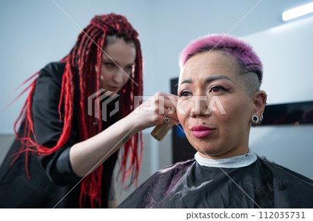 The hairdresser shaves the temple of a female client. Rear view of a woman with short pink hair in a barbershop. The hairdresser shaves the temple of a female client. Rear view of a woman with short pink hair in a barbershop. 112035731