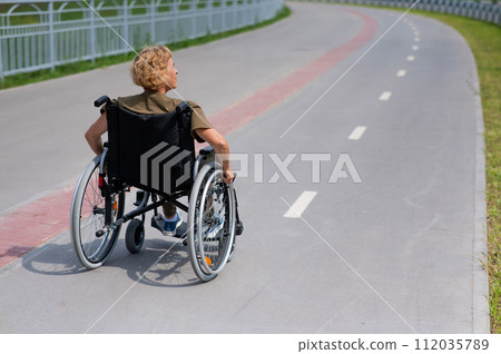 Rear view of an elderly woman in a wheelchair riding on a bike path. Rear view of an elderly woman in a wheelchair riding on a bike path. 112035789