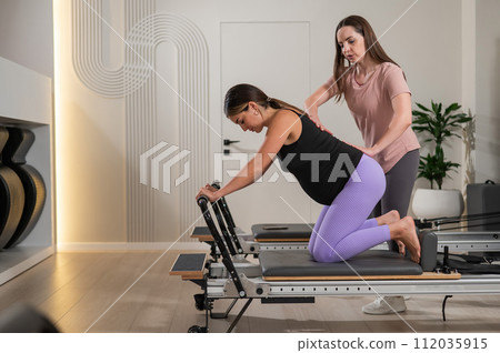 A pregnant woman works out on a reformer exercise machine with a personal trainer. A pregnant woman works out on a reformer exercise machine with a personal trainer. 112035915