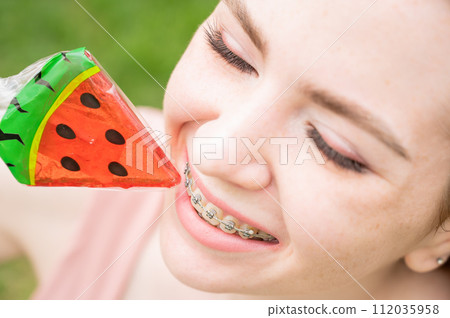 Beautiful young woman with braces on her teeth eats a watermelon-shaped lollipop outdoors. Beautiful young woman with braces on her teeth eats a watermelon-shaped lollipop outdoors. 112035958