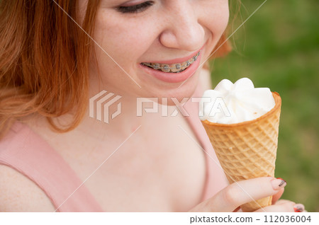 Young beautiful red-haired woman smiling with braces and eating an ice cream cone outdoors. Young beautiful red-haired woman smiling with braces and eating an ice cream cone outdoors. 112036004