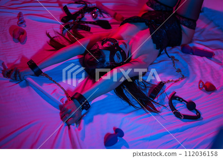 A woman in a leather mask and handcuffs lies on a bed in a blue-red light. Sex toys collection. A woman in a leather mask and handcuffs lies on a bed in a blue-red light. Sex toys collection. 112036158