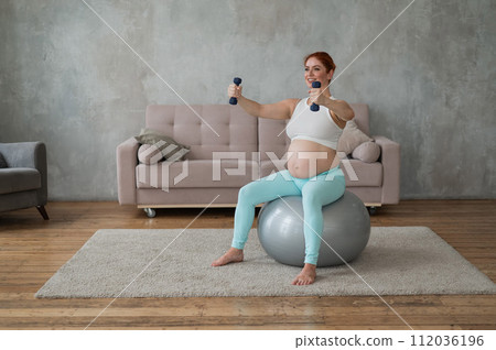 Pregnant woman doing exercises with dumbbells while sitting on a fitness ball at home. Pregnant woman doing exercises with dumbbells while sitting on a fitness ball at home. 112036196