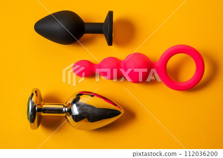 A set of three alternative sex devices of various shapes and sizes on an orange background. Silicone and metal plugs and anal beads. A set of three alternative sex devices of various shapes and sizes on an orange background. Silicone and metal plugs and anal beads. 112036200