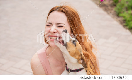 Dog jack russell terrier licks the owner in the face outdoors. Girl with braces on her teeth.  112036203