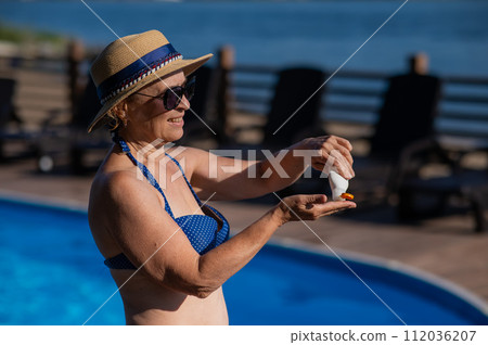 Portrait of an old woman in a straw hat, sunglasses and a swimsuit applying sunscreen to her skin while relaxing by the pool.  112036207