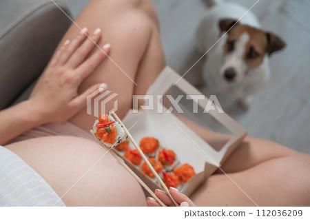 A pregnant woman sits on the sofa and eats rolls. Jack Russell Terrier dog sits on the floor and begs for food from its owner.  112036209