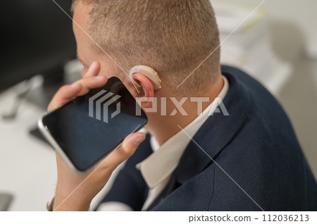 Caucasian man with a hearing aid talking on the phone on speakerphone.  112036213