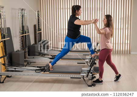 Overweight caucasian woman doing pilates exercises on reformer with personal trainer.  112036247