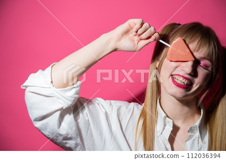 Portrait of a young woman with braces and bright makeup eating a lollipop on a pink background. Portrait of a young woman with braces and bright makeup eating a lollipop on a pink background. 112036394