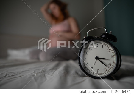 Caucasian pregnant woman sits on the bed and suffers from insomnia. Alarm clock in the foreground.  112036425