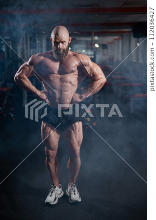 Muscular bald man posing in shorts. Bodybuilder showing off his shape in the gym. Muscular bald man posing in shorts. Bodybuilder showing off his shape in the gym. 112036427