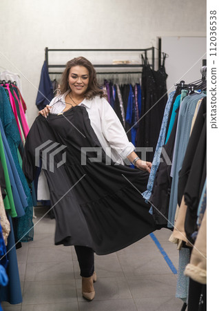 A fat woman in a plus size store tries on a black evening dress. Vertical photo.  112036538