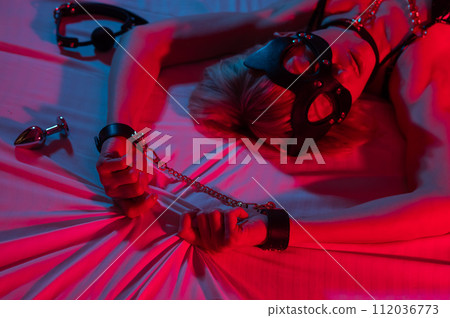 Sexy blonde woman in a leather mask and handcuffs and a collar lies in the bedroom in a red-blue neon light. Role-playing games. Sexy blonde woman in a leather mask and handcuffs and a collar lies in the bedroom in a red-blue neon light. Role-playing games. 112036773