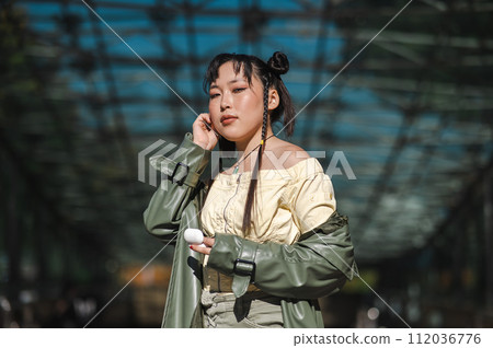 A beautiful Asian woman in shorts and a green leather coat comes out of the subway and uses wireless headphones.  112036776